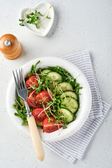 Fresh salad with tomato, cucumber, vegetables, microgreen radishes in white plate on grey stone background. View from above. Concept vegan and healthy eating.