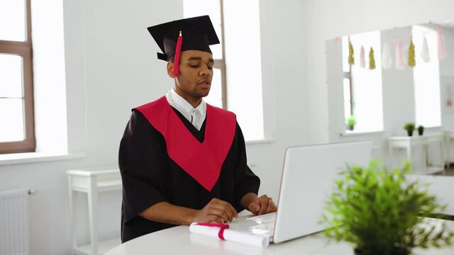 A black student sits at a laptop in a gown and square cap. Virtual graduation and convocation ceremony at home while quarantine
