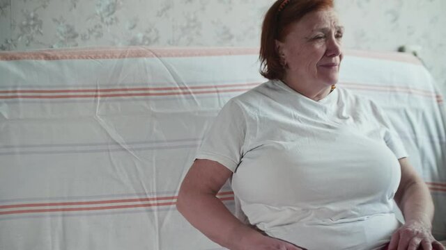 Old woman tries to stand up from sofa. She feel pain and sit down stroking her back. Old people with bad health.
