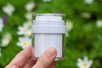 A white jar of cosmetics in your hand on a background of greenery and white flowers