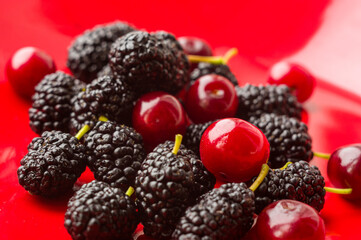 Ripe large black mulberries and red cherries on a red plate, close-up.