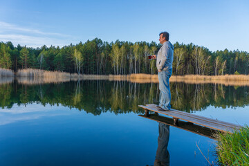 Fototapeta na wymiar Happy man stands on a wooden pier with glass cup tea near spring forest on a calm lake. Nature and travel concept