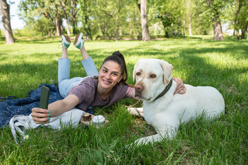 Young beautiful woman lying in the park with her pet dog a golden Labrador Retriever and takes a selfie photo with her smartphone while having fun and enjoying a beautiful sunny day