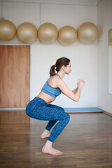 A woman in sportswear is engaged in Pilates, doing squats in the gym, side view