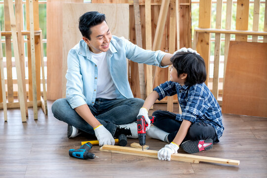 Asian father touch head of his son during drill the timber in home workplace of carpentering with happy emotion. Asian family concept to stay at home and enjoy good relationship hobby together.