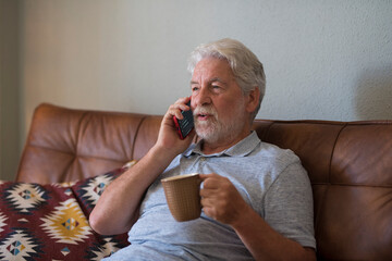 Old senior man at home speaking with modern phone and drinking coffee or tea sitting on the sofa...
