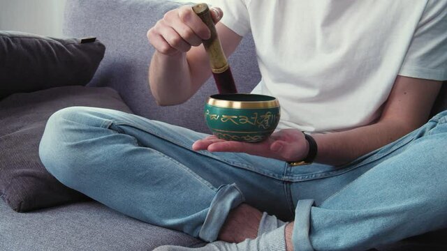 Young man using tibetan singing bell bowl at home living room, carrying about mental health. Letters mean tibetan mantra: Om Mani Padme Hum. English translation: Praise To The Jewel in The Lotus