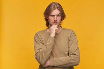 Young man, thoughtful guy with blond hair, beard and mustache. Wearing beige sweater. Touching his chin and lifts an eyebrow. Watching at the camera isolated over yellow background