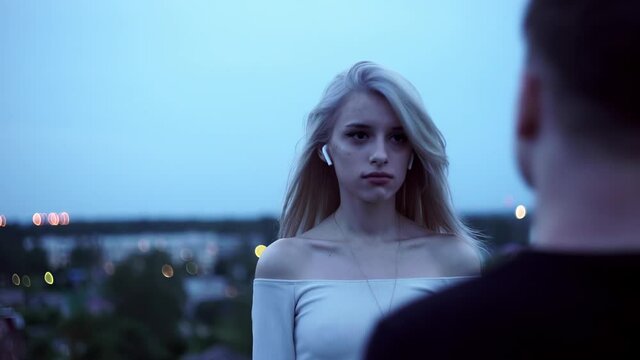 The girl sadly looks at her boyfriend standing in front of him on the roof in the evening. Emotions on the face. Behind them, you can see the city lights.