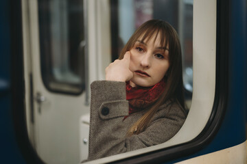 Fototapeta na wymiar young caucasian woman with long brown hair wearing elegant classy coat and a scarf sitting alone in subway carriage and distantly looking out window. Image with selective focus and toning
