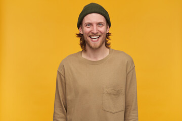 Young man, cheerful guy with blond hair, beard and mustache. Wearing green beanie and beige sweater. Watching at the camera and broadly smiling, isolated over yellow background