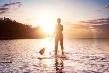 Fototapeta premium Adventurous girl on a paddle board is paddeling in the Pacific West Coast Ocean. Sunset Sky Art Render. Taken near Spanish Banks, Vancouver, British Columbia, Canada.