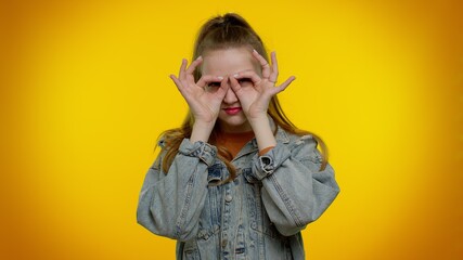 Nosy curious teen stylish girl closing eyes with hand and spying through fingers, hiding and peeping, binocular gesture, exploring way, seeking something in distance. Young woman on yellow background