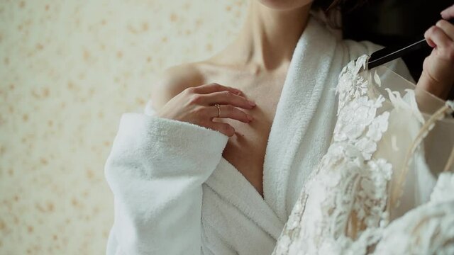 Caucasian woman in terry robe holds a wedding dress on a hanger in her hand. She gently strokes her bare shoulder in hotel room 