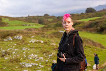 Young woman with pink hair, punk aesthetics and androgynous features holding a camera in the middle of a natural environment. Alternative and rebellious style traveler.