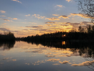Dramatic sunset sky, clouds reflect in still water surface of forest lake, bare trees silhouetted on horizon