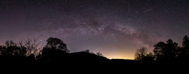 A panoramic view of the Milky Way arching over Aira Farce & the trees & mountains next to Ullswater