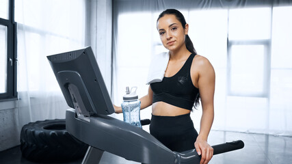 sweaty sportswoman with towel on treadmill looking at camera near sports bottle with water in gym.