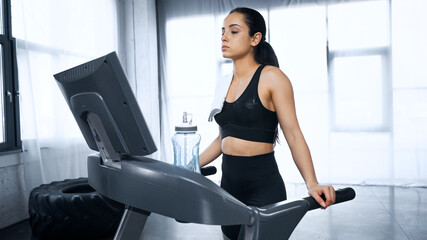 Fototapeta premium sporty woman with towel walking on treadmill near sports bottle with water in gym.