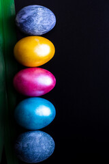 Multi-colored painted easter eggs on top of each other are laid out on a black background with a dark green leaf on the side and with a place for the inscription on the right