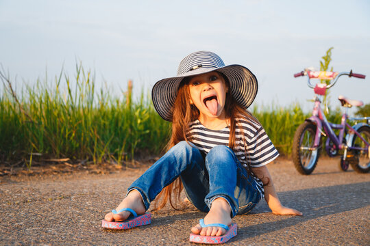 Cute Little Girl Showing Tongue Out In Big Sun Hat Sitting On Road With Her Bicycle Behind Her