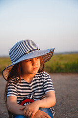 Close up portrait of little sad girl in blue and white striped sun hat and shirt sitting alone at the road