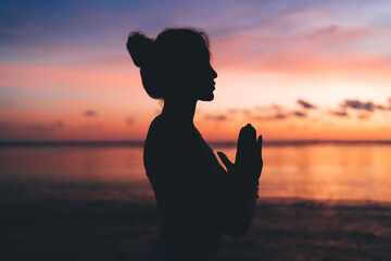 Side view of female silhouette keep praying during mindfulness meditation at seashore beach with colorful sky, slim woman in Namaste relaxing during evening physical recreation at coastline