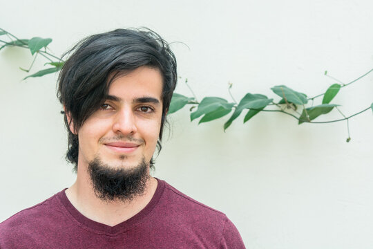Portrait of a young caucasian black haired man with beard and red shirt looking at camera and smiling naturally, confident and friendly. White background and climbing plant with leaves behind - Powered by Adobe
