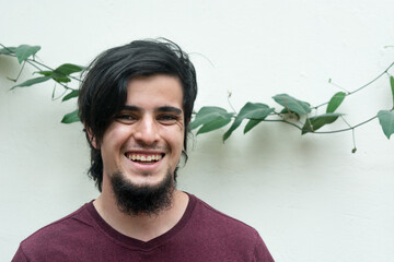 Portrait of young caucasian black haired man with beard and red shirt looking at camera, laughing and smiling naturally, confident and friendly. White background and climbing plant with leaves behind