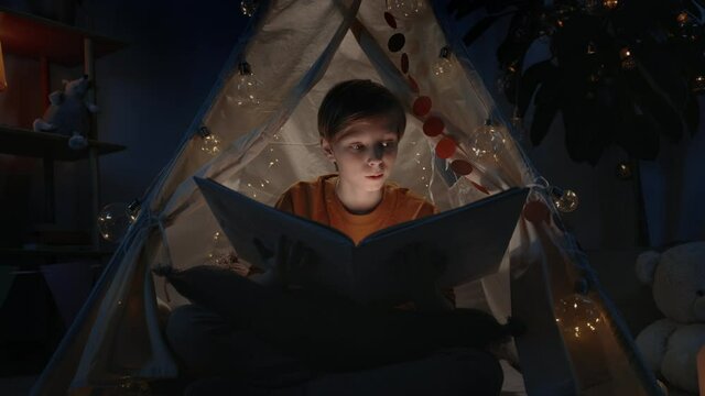 Excited boy with blond hair in makeshift hut opening book at home in evening. Teenager sitting on floor while reading book with flashlight. Concept of leisure and free time.