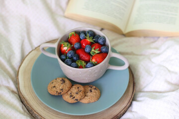 Bowl of blueberries and strawberries, chocolate chip cookies and open book on a bed. Selective focus.