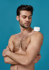 Fototapeta premium Studio shot of naked brunette guy looking at white jar of effective cream on his shoulder, posing with arms crossed isolated over blue background