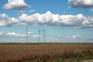 Fototapeta na wymiar Nature and industrialisation: high voltage electric wire in the field with blue sky and white clouds; color photo.