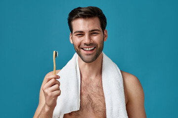 Beauty portrait of happy guy with towel around his neck smiling at camera, holding toothbrush while...