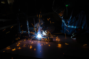 A man welder with construction gloves and a welding mask is welded with a welding machine metal in workshop.