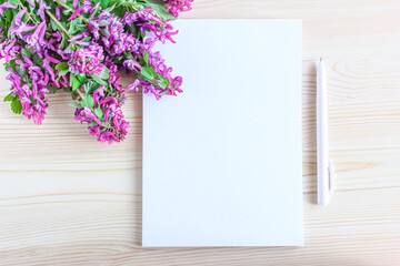Blank notebook, pen and pink flowers with green leaves on a wooden background, top view. White sheet with copy space.