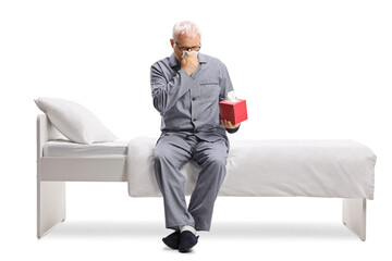 Man in pajamas sitting on a bed and blowing his nose with paper tissues