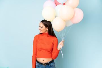 Young woman catching many balloons isolated on blue background looking to the side