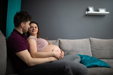 A married couple looks at each other with great joy while sitting on the sofa in the living room. The woman is pregnant.