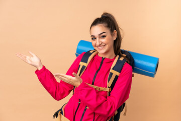 Young mountaineer girl with a big backpacker isolated on beige background extending hands to the side for inviting to come