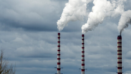 Industrial smoke from chimney on dramatic sky background. View of high chimney pipes with grey smoke from coal power plant. Production of electricity with fossil fuel. Wind blowing pollution.