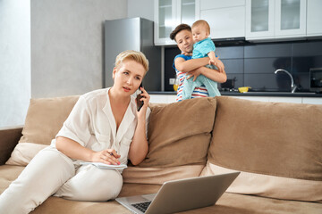 Busy mother talking on mobile and working on modern laptop while her children playing together. Woman doing remote work at home during maternity leave.