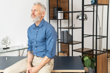 Tranquil contemporary middle aged businessman in smart casual wear leaned on the desk and looks away, inspired and serene mature business owner at the office, confident grey-haired man in relaxed pose