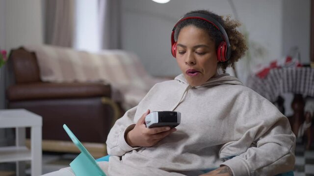 Middle shot of dissatisfied African American woman printing Remote working with label maker sitting on bag chair in home office. Portrait of sad young worker with tablet indoors