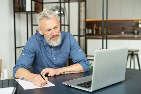 Modern middle aged bearded tattooed man working with a laptop in home office, senior grey hair male in smart casual shirt watching webinar and writes down, takes notes with a pen, planning schedule