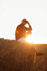handsome young man in sunglasses, casual black sweatshirt and white short, in summer field outdoor, sitting on hay bale, haystack on sunset