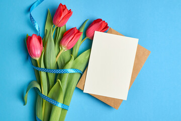 Greeting card mockup with copy space, envelope and red tulips bouquet with ribbon on blue paper background