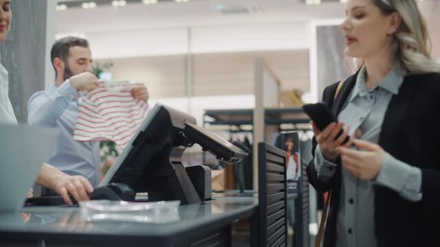 Clothing Store: Young Woman At Counter Buys Clothes from Friendly Retail Sales assistant, Paying with Contactless NFC Smartphone Touching Terminal. Trendy Fashion Shop with of Designer Brands.