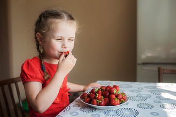 Cute little girl in a red t-shirt sits at the table and eats strawberries from a plate
