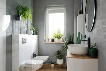 Interior of stylish bathroom with ceramic washbasin, round mirror, green plants, window, grey wall....